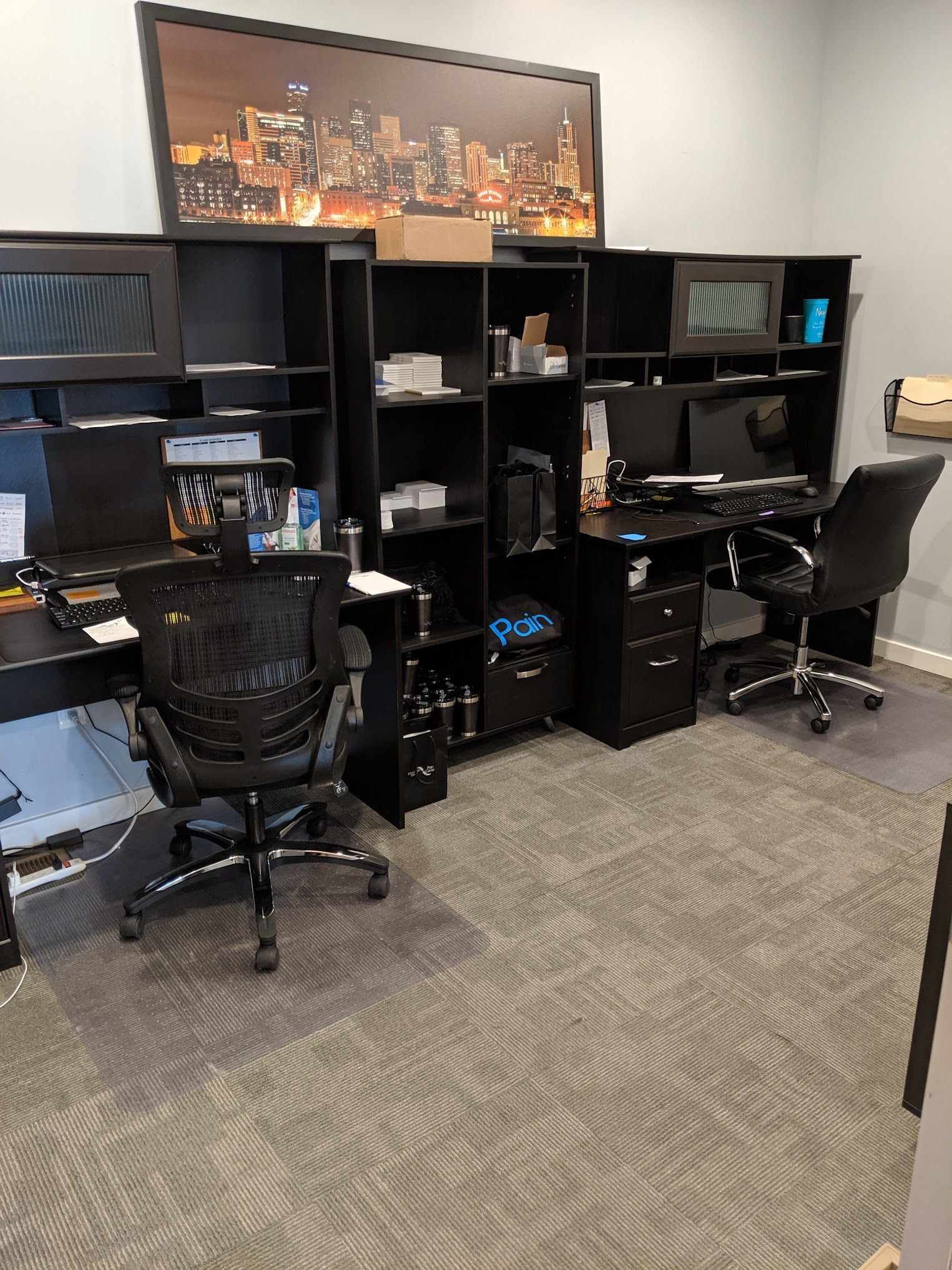 Two black desks with monitors, chairs, and storage against a wall with a city skyline photo.