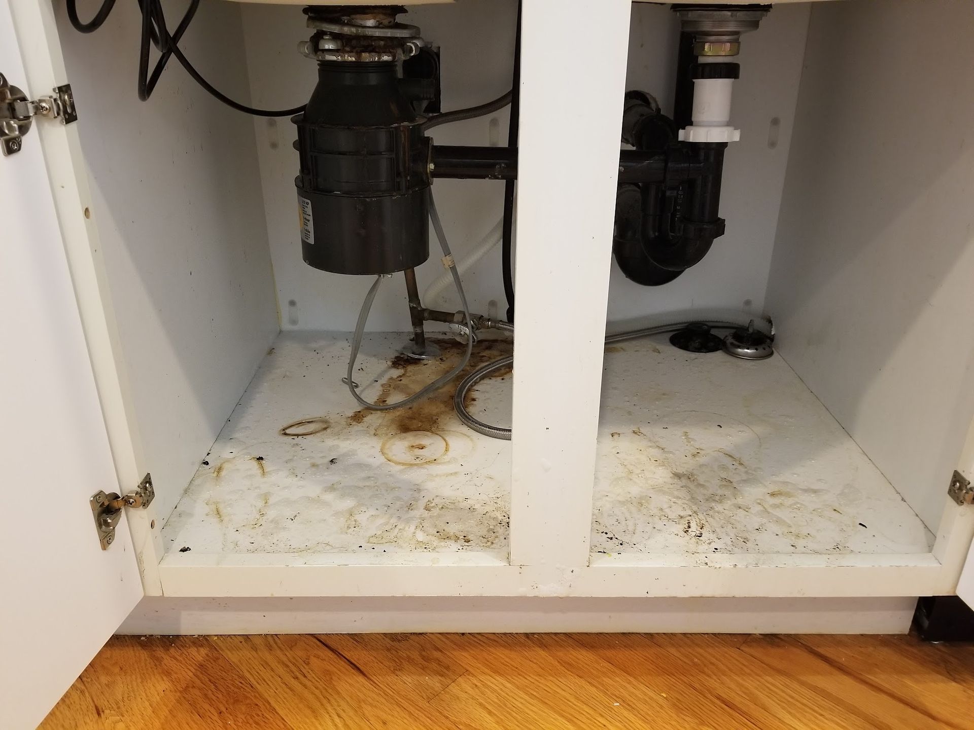 Under kitchen sink, cabinet open, plumbing and garbage disposal visible, stained floor.