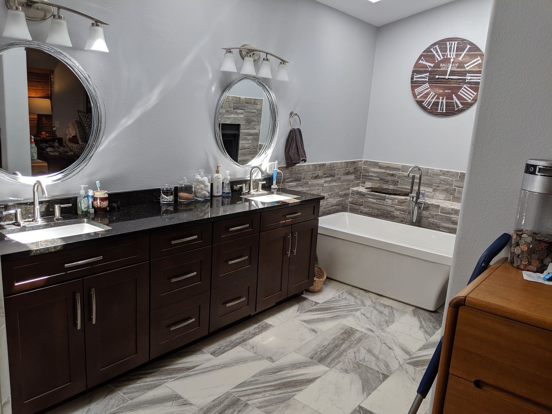 Bathroom with dark cabinets, large mirrors, stone wall, tub, and a clock.