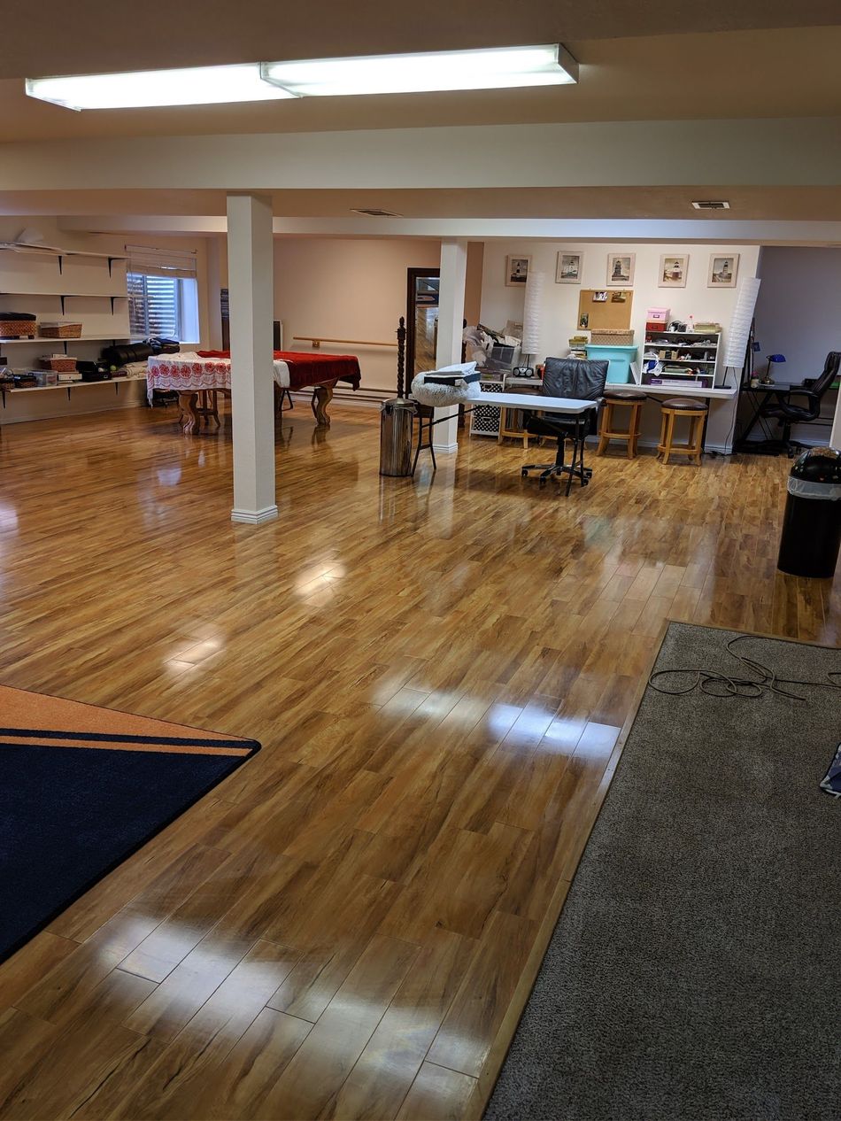 A large, open room with wood flooring and work tables. White support beams, bright overhead lights.