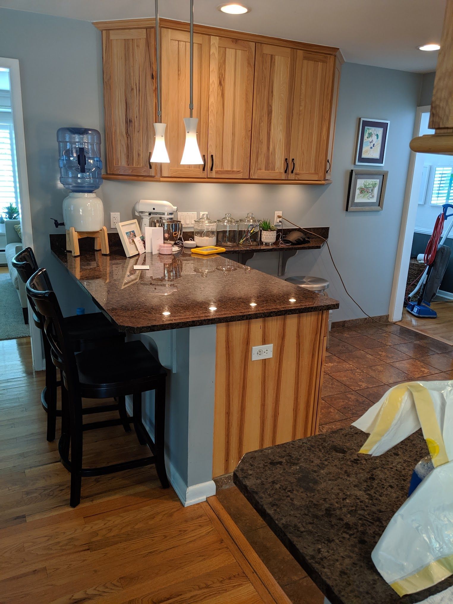 Kitchen with granite countertop, wooden cabinets, and bar stools. Water cooler and hanging lights are visible.