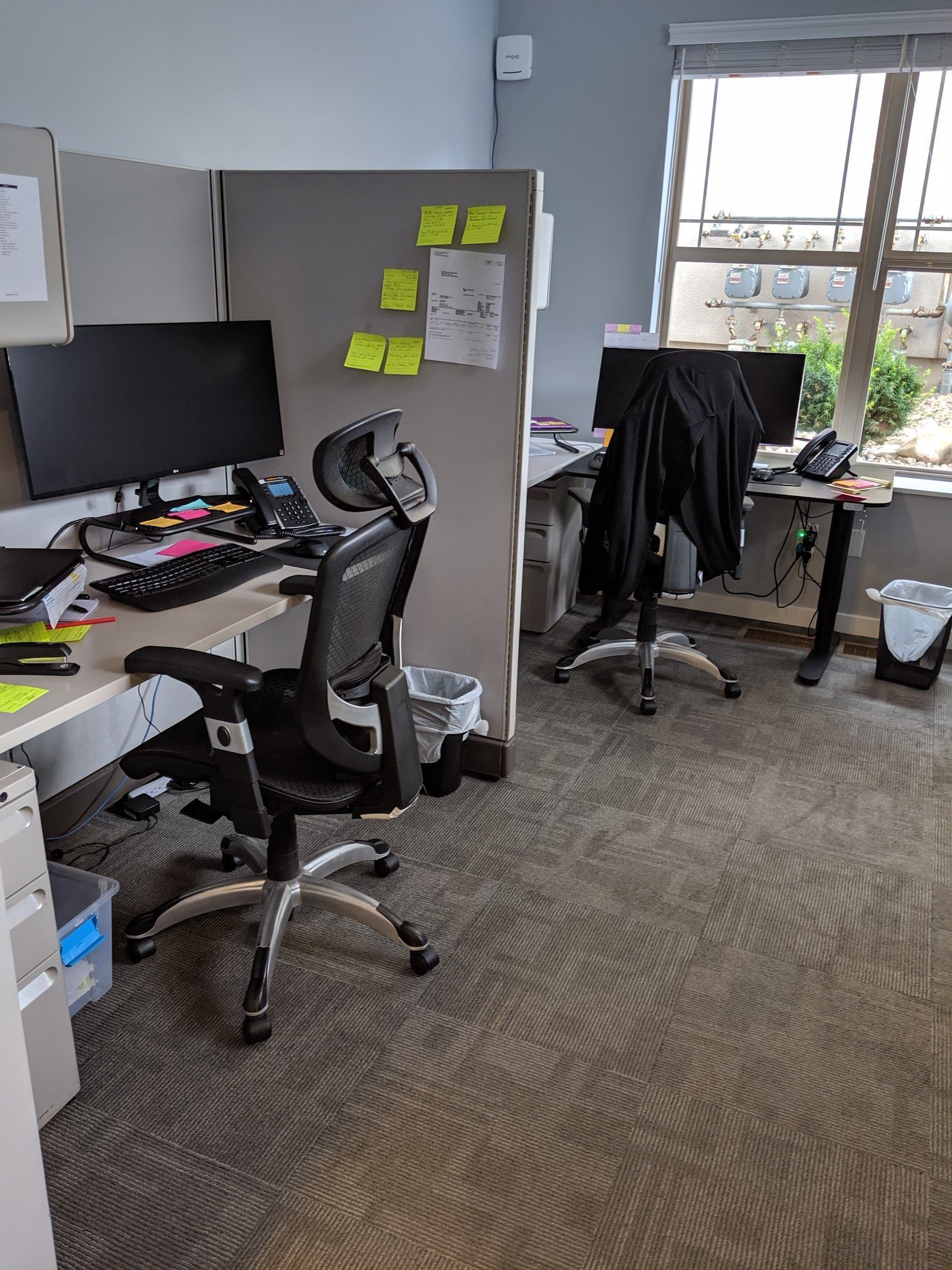 Office cubicles with desks, chairs, and monitors. One chair is in front, a black jacket hangs on another.