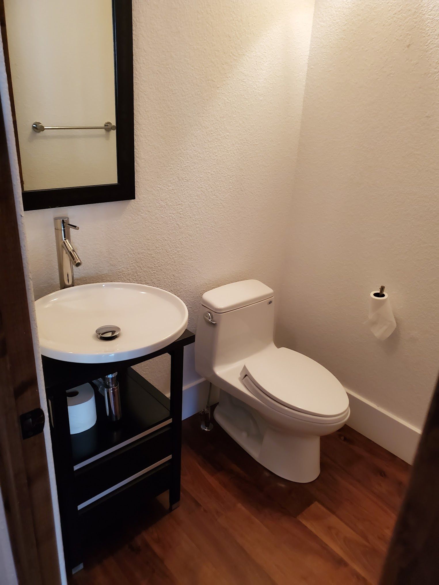 Small bathroom with a white toilet, round sink, dark wood floor, and a black-framed mirror.