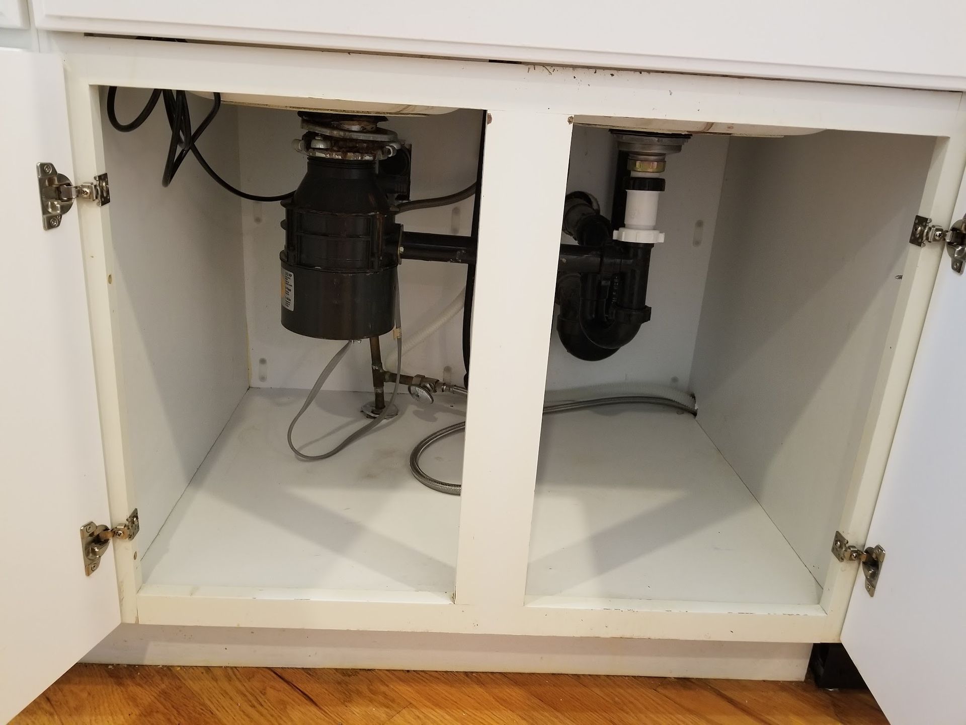 Under-sink cabinet with garbage disposal and plumbing visible. White cabinet interior, hardwood floor.