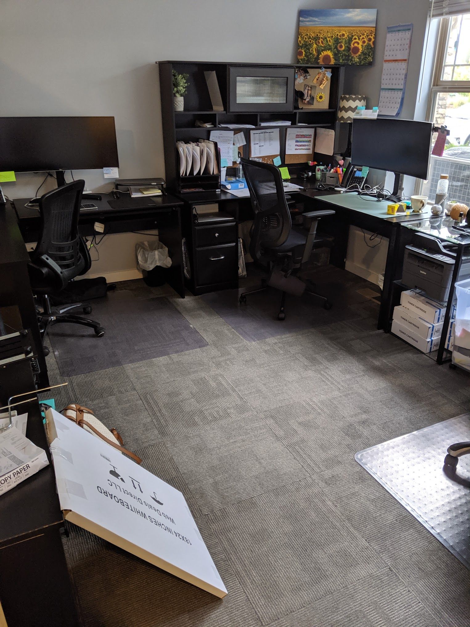 Office space with multiple desks, computers, chairs, and assorted supplies. The carpet has a grey swirling pattern.