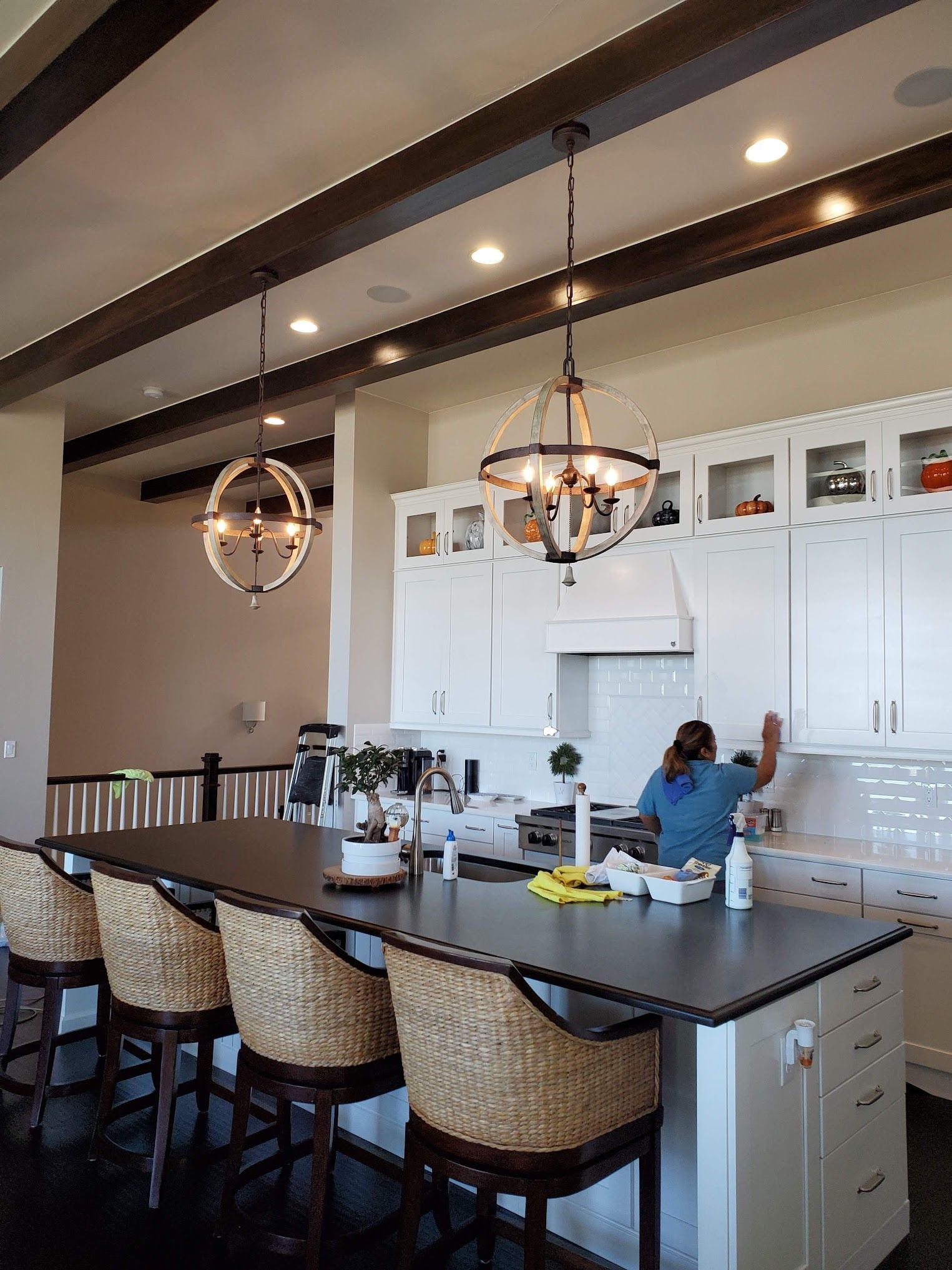Kitchen with island, white cabinets, and dark wood beams. A person cleans the countertop.