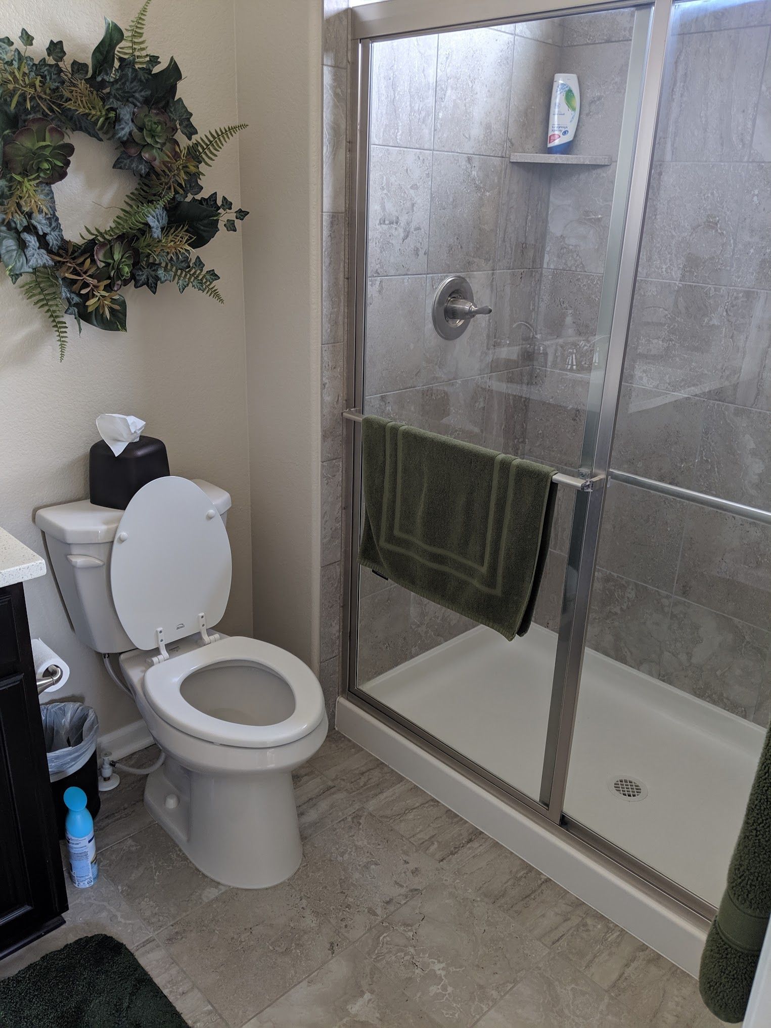 Bathroom with toilet, shower, and dark green towel. Gray tile and a decorative wreath on the wall.