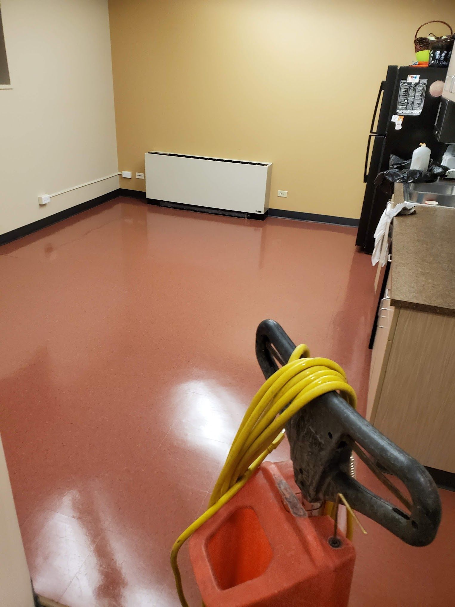 A red floor in a room with a cleaning machine. Yellow cord. Beige walls and a white heater.