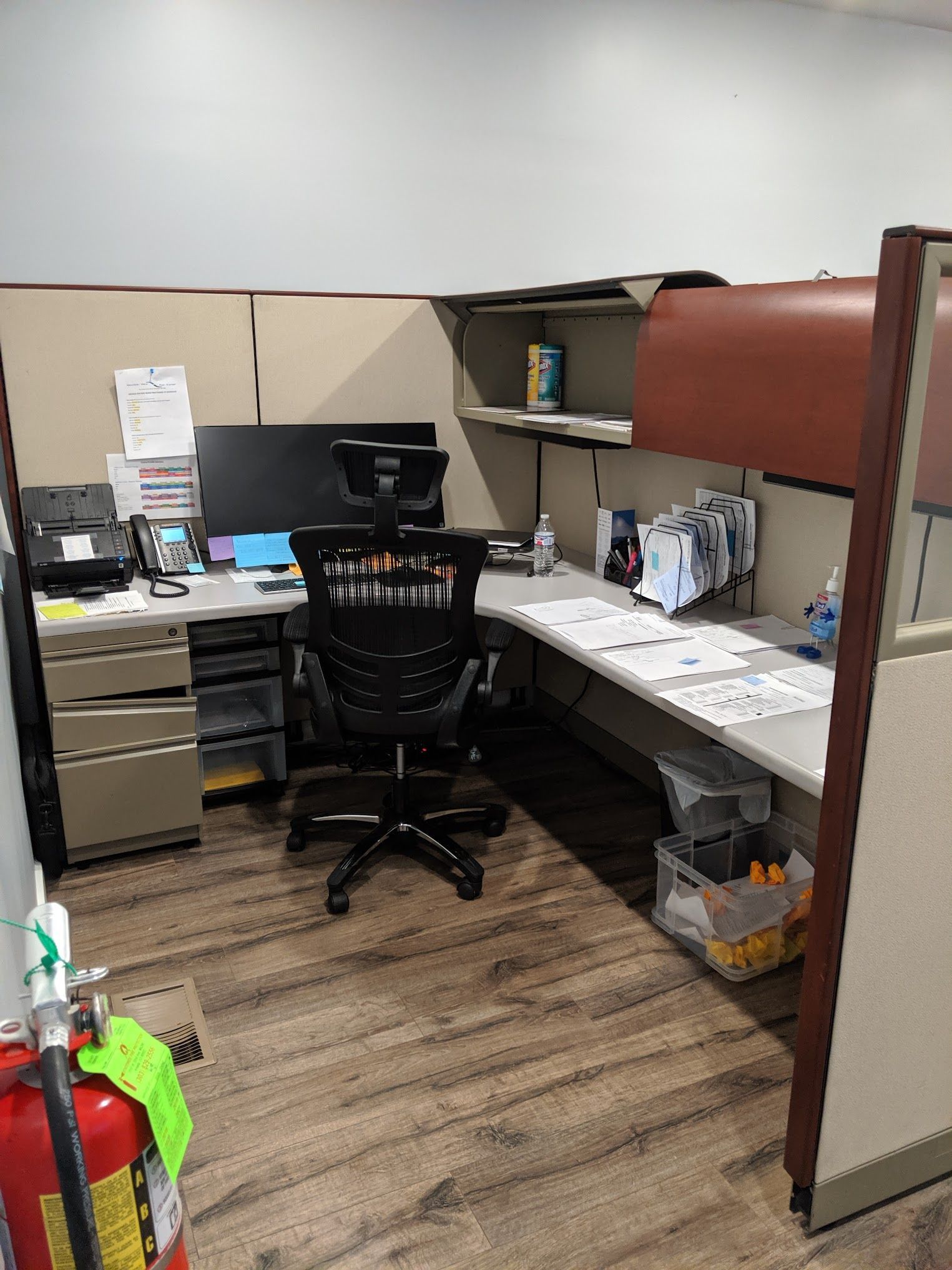 Office cubicle with desk, computer, chair, and papers. Includes a fire extinguisher in the foreground.