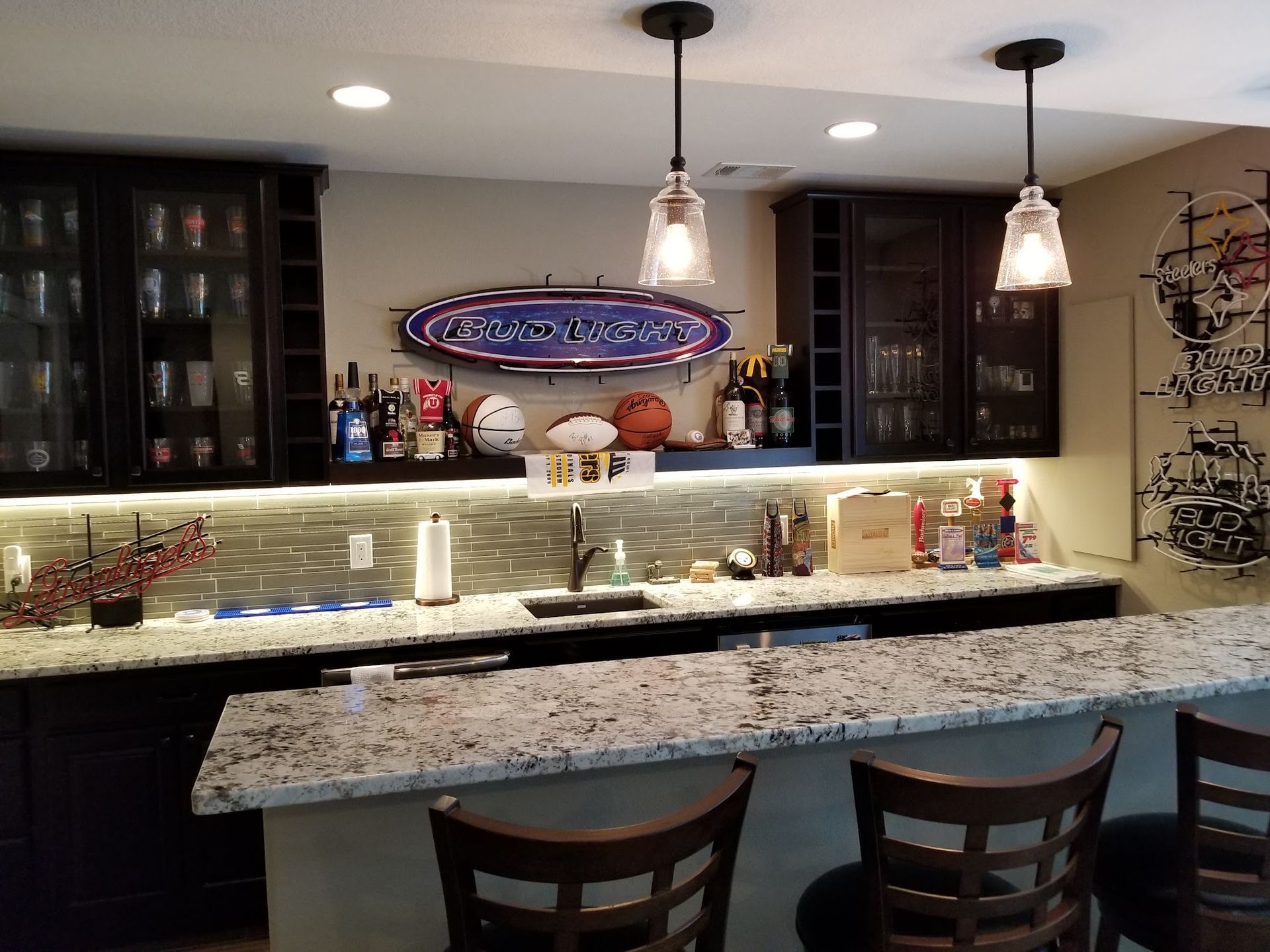 Basement bar with granite countertop, dark cabinets, and sports memorabilia. Two pendant lights illuminate the bar area.