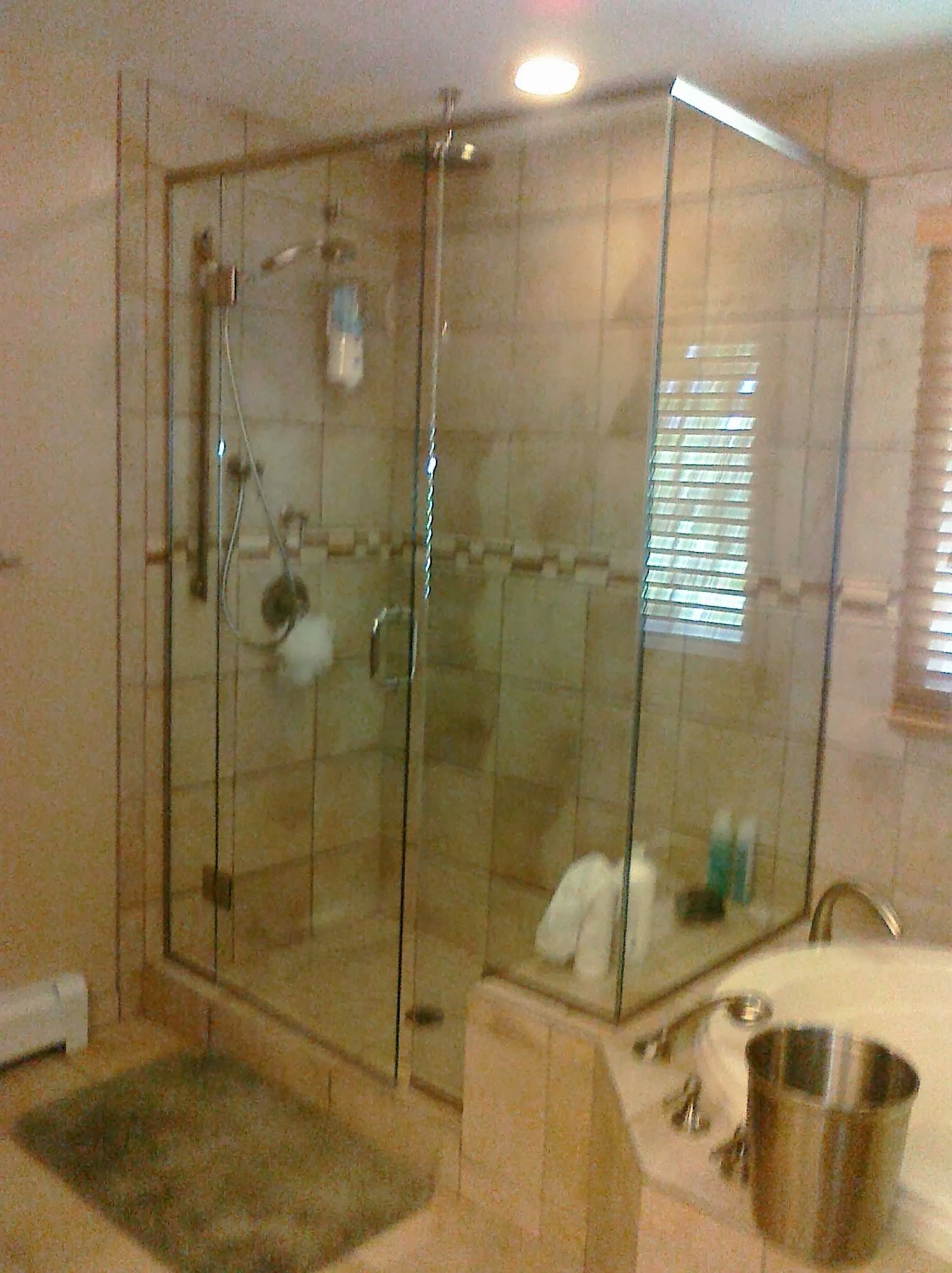 Glass shower enclosure in a beige-tiled bathroom, with a tub and window visible.