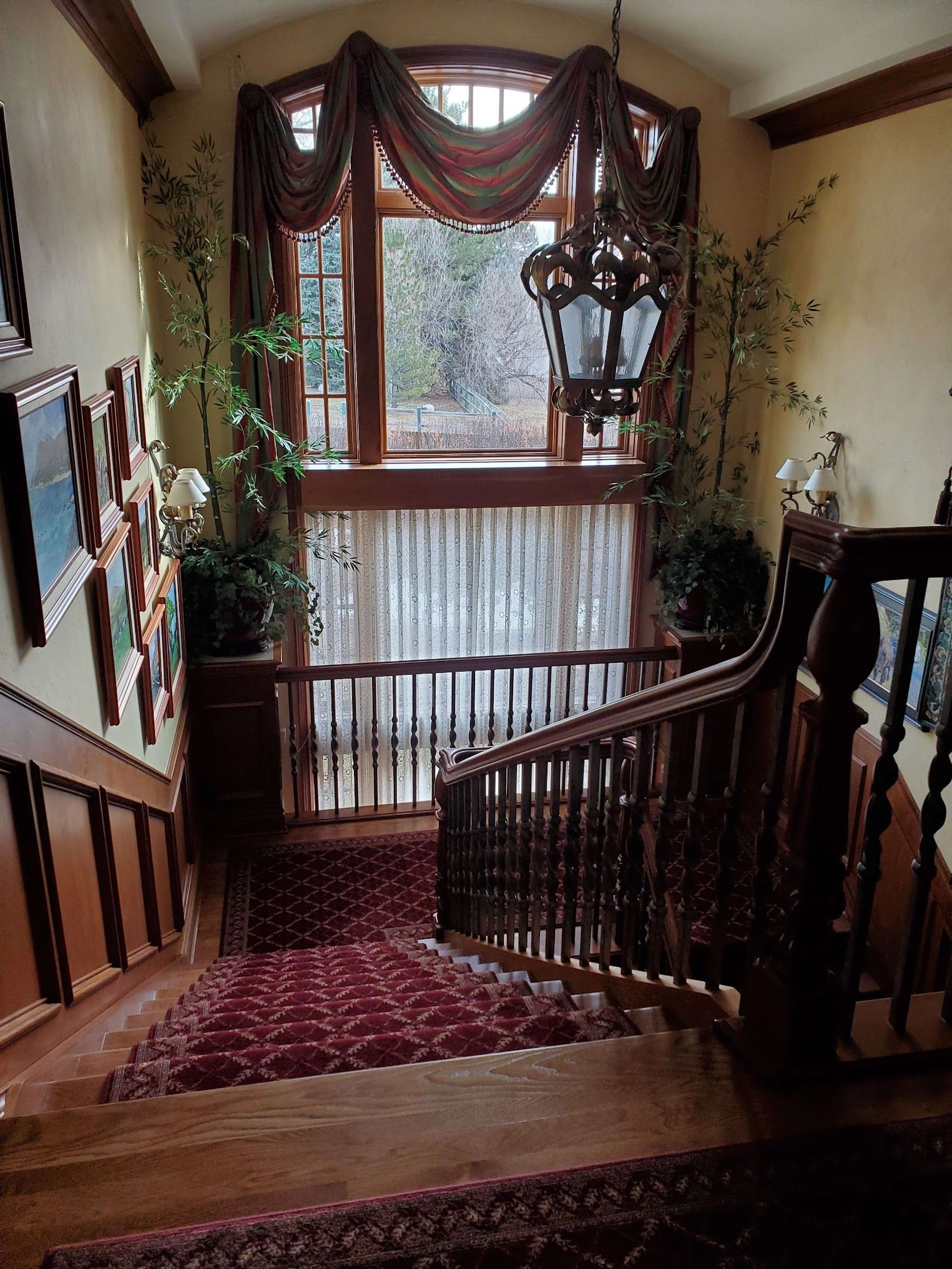 Wooden staircase with red carpet, ornate railing, and large window with draping.