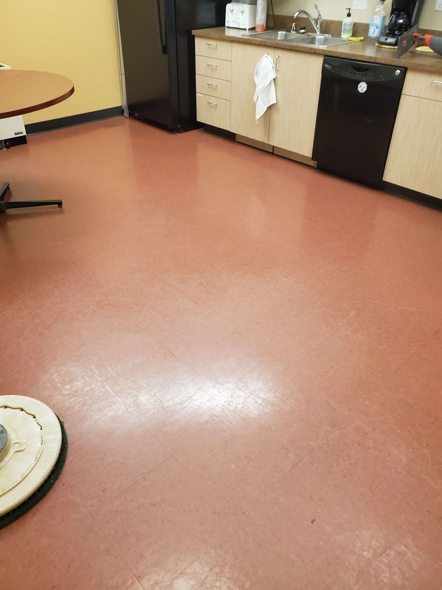Pink-red floor of a kitchen area with a black dishwasher and cabinets. A floor cleaning machine is on the floor.