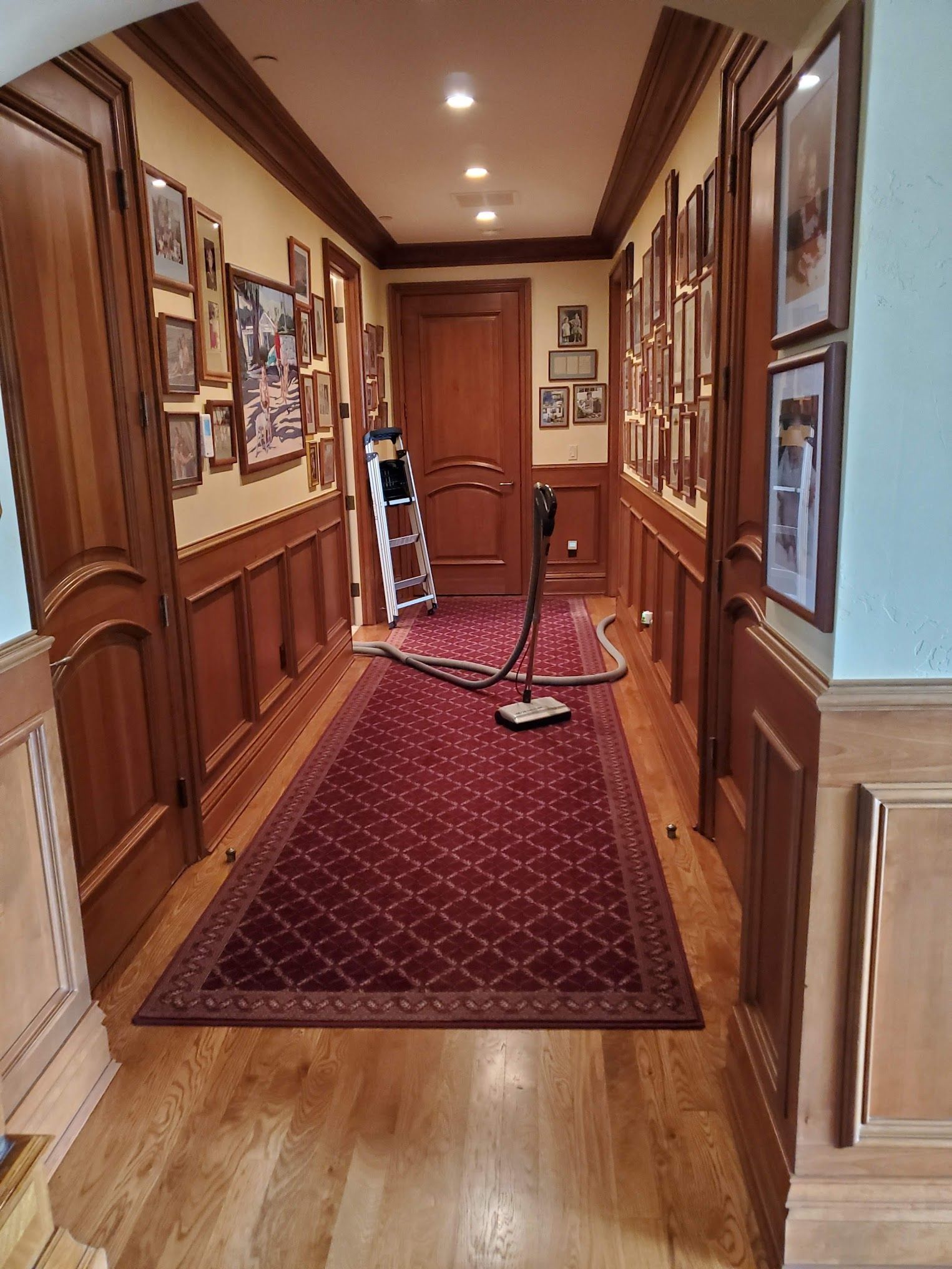 Long hallway with red carpet, wood paneling, and framed artwork on the walls. A vacuum cleaner sits on the rug near a door.