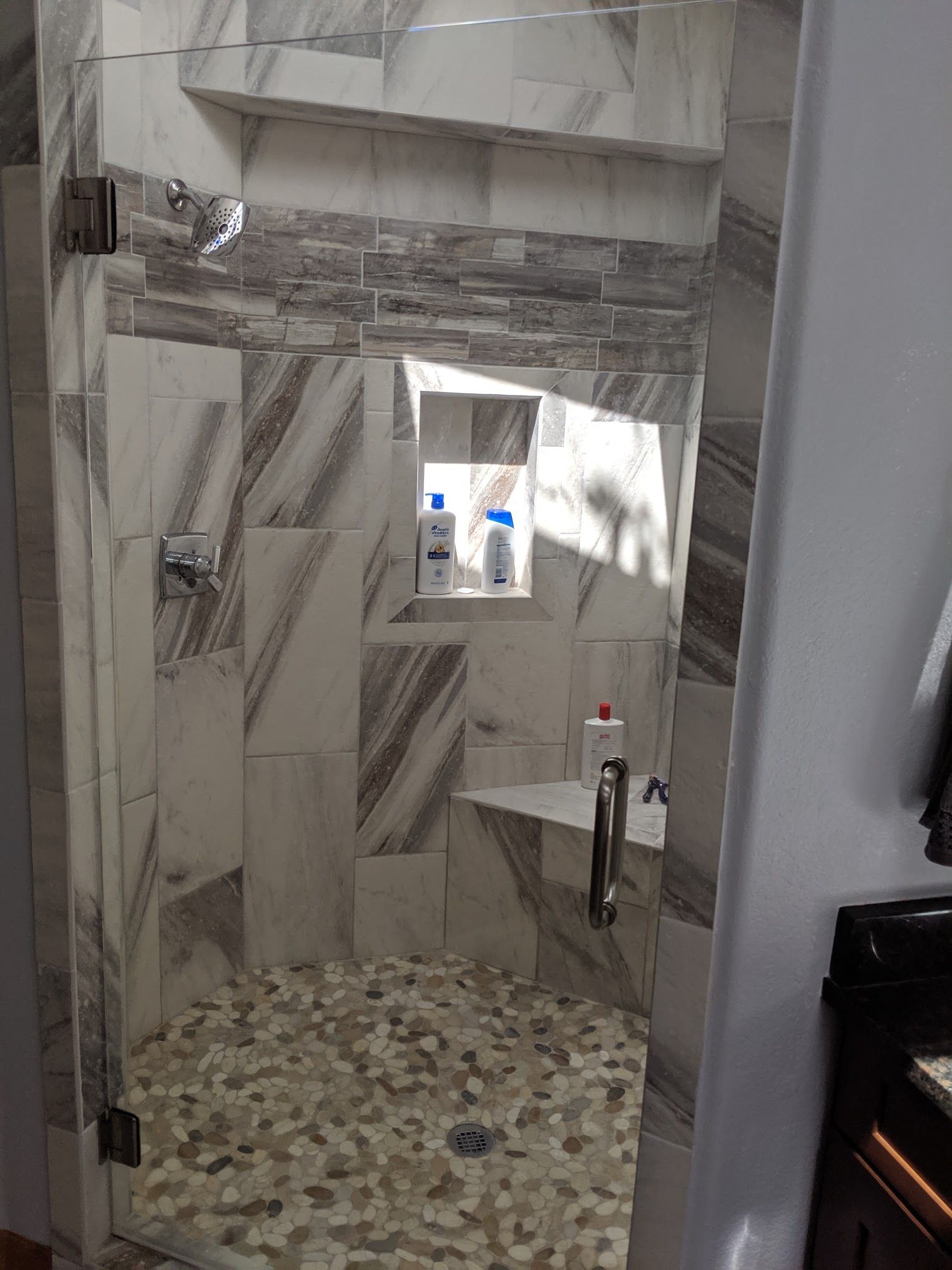 A tiled shower with a pebble floor and glass doors, featuring a built-in shelf with soap bottles.