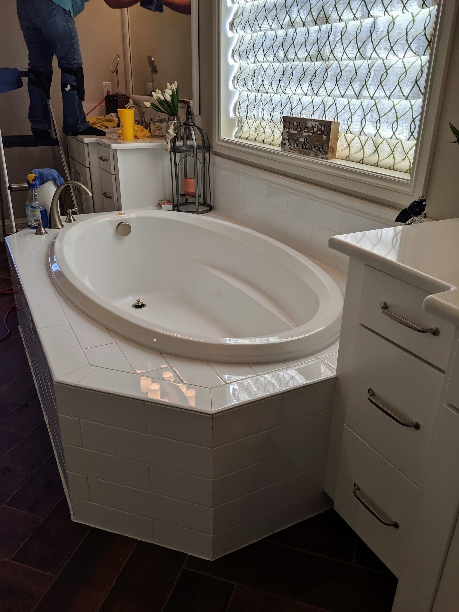 Oval bathtub set in a white tiled platform near a window. A person stands on a ladder nearby.