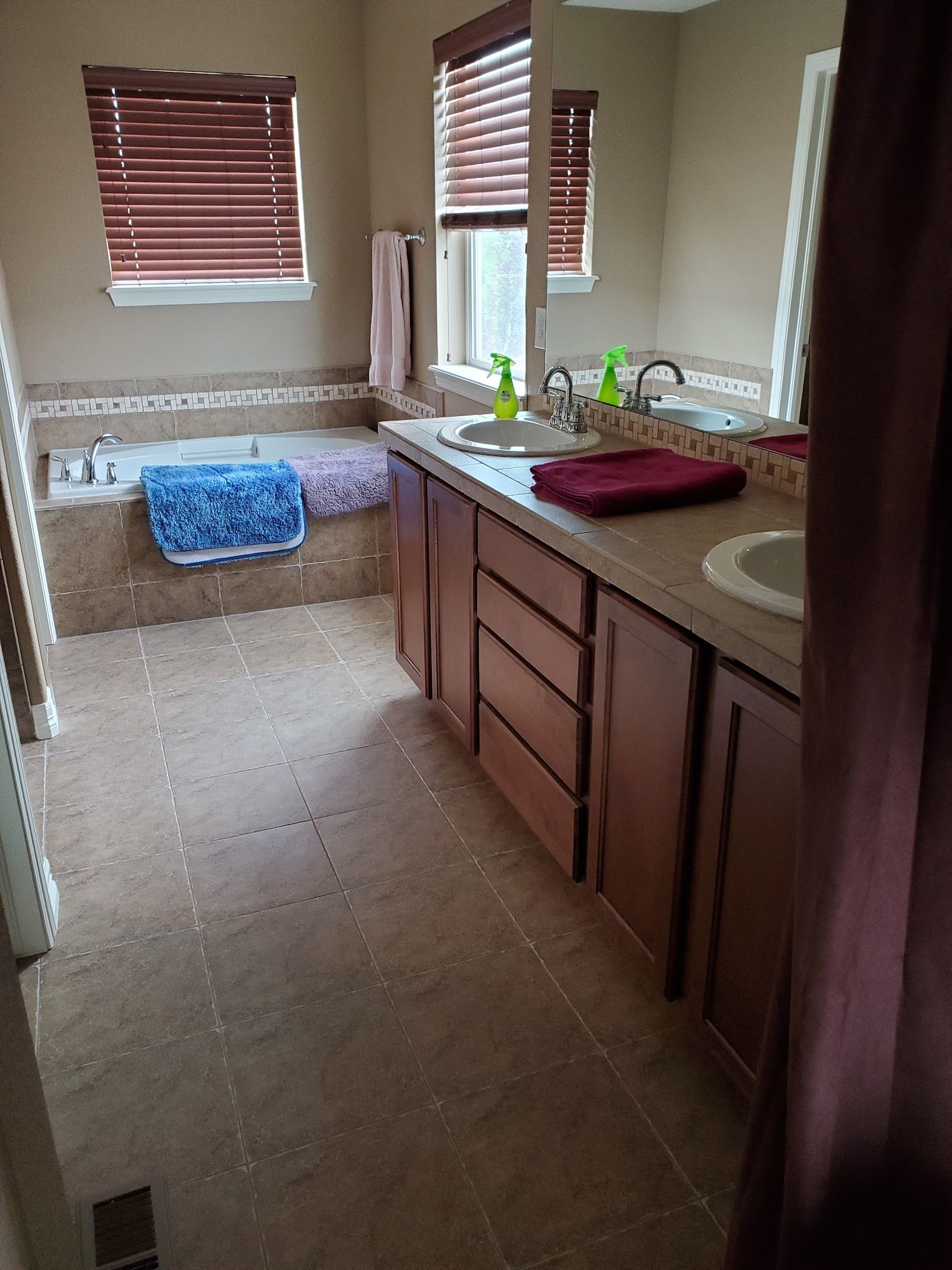 Bathroom with a built-in tub, double vanity, and brown cabinetry. Brown tile flooring and wooden blinds.