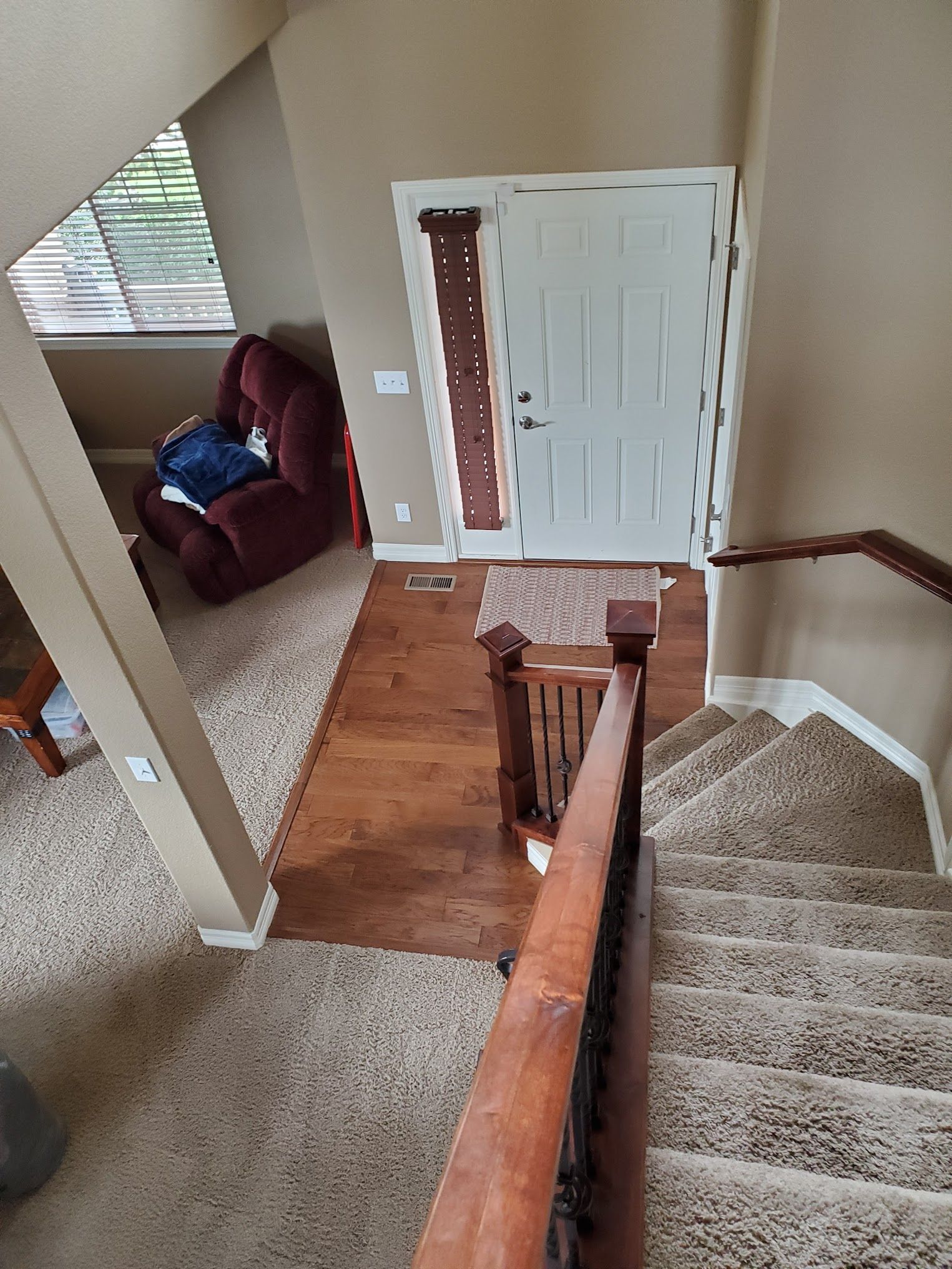 Entryway with stairs, door, and seating area. Brown handrail, hardwood floor, tan carpet, and burgundy chair.