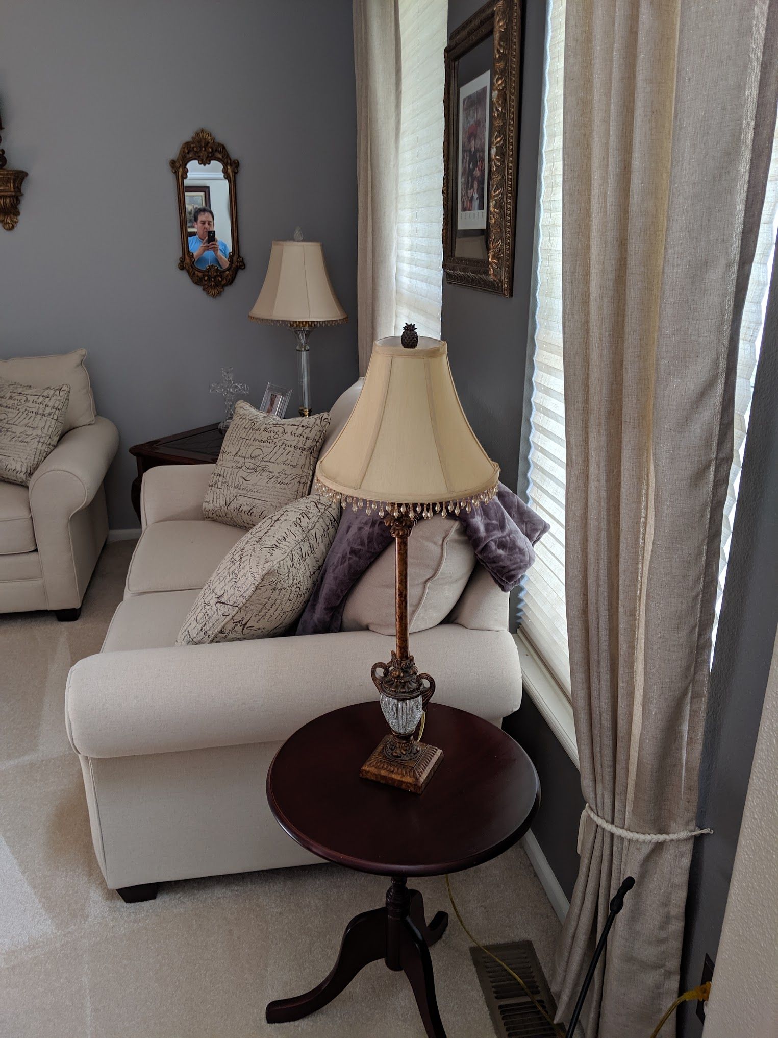 Living room scene: cream-colored couch, side table with lamp, window, and decorative mirror on gray wall.