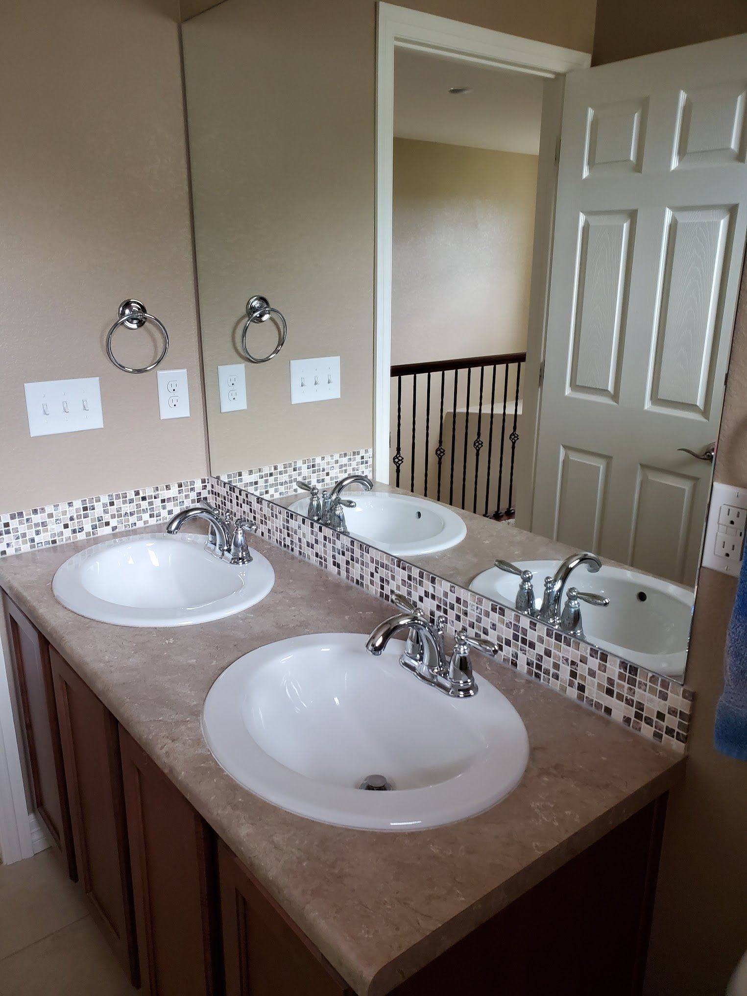 Bathroom with double sinks, large mirror, brown cabinets, and mosaic tile backsplash. Doorway to stairs visible.