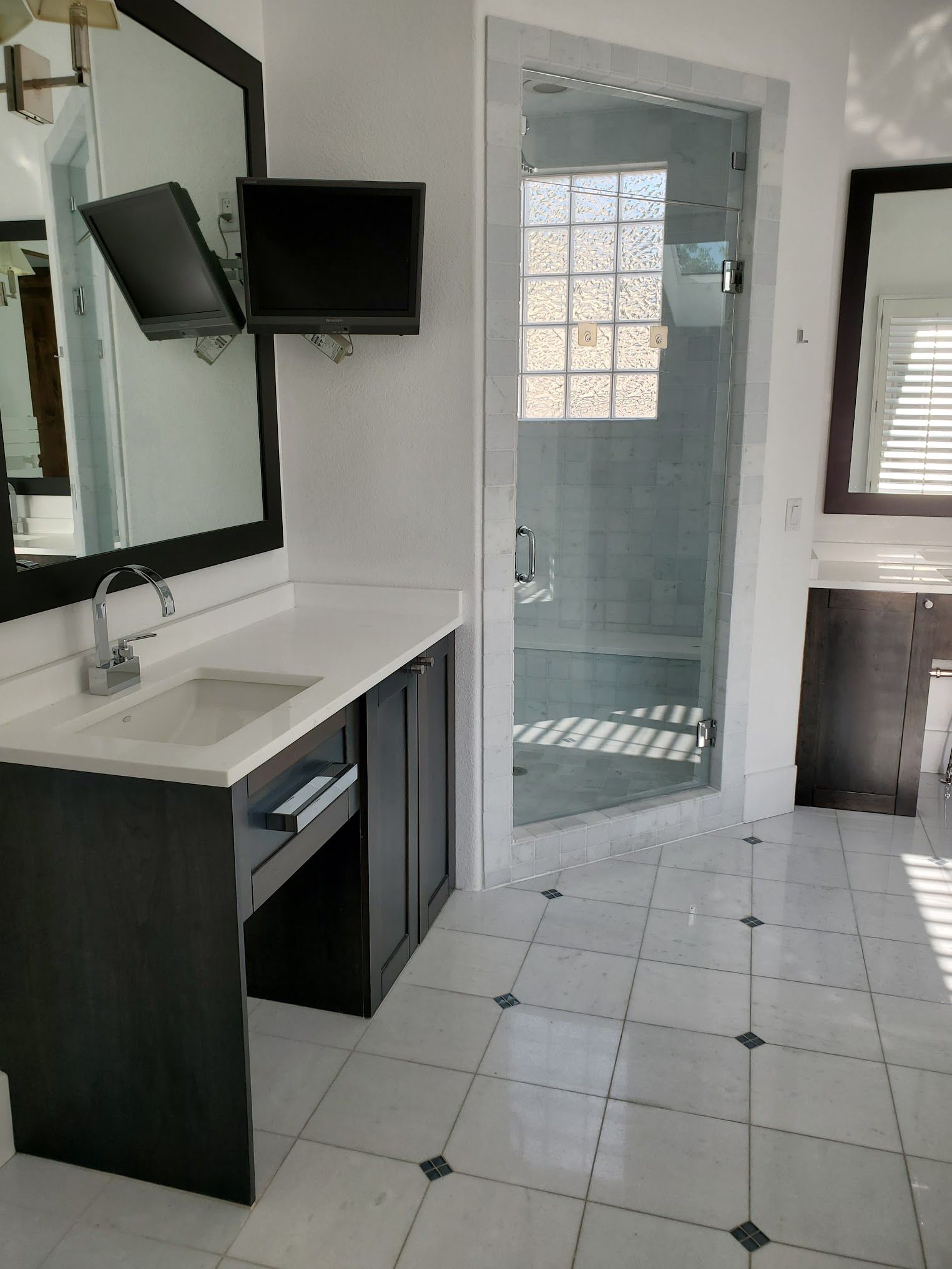 Bathroom with dark vanity, large mirrors, glass shower, and a wall-mounted television. White and gray color scheme.