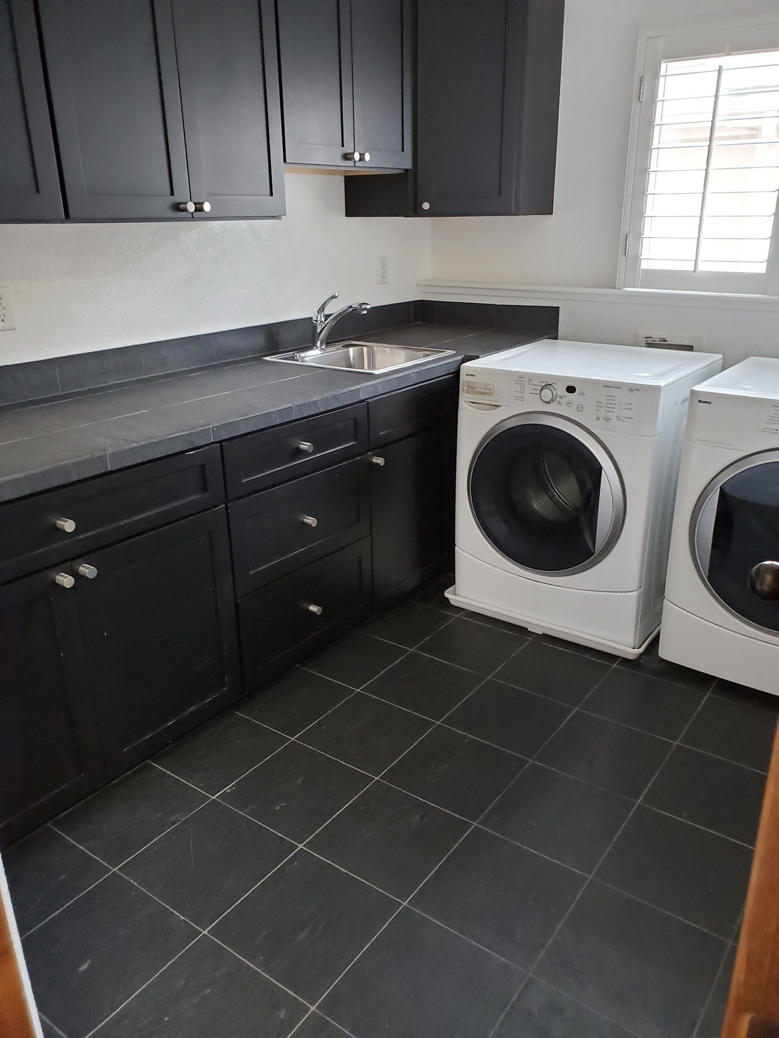 Laundry room with black cabinets, sink, and white washer/dryer. Black tiled floor and window.