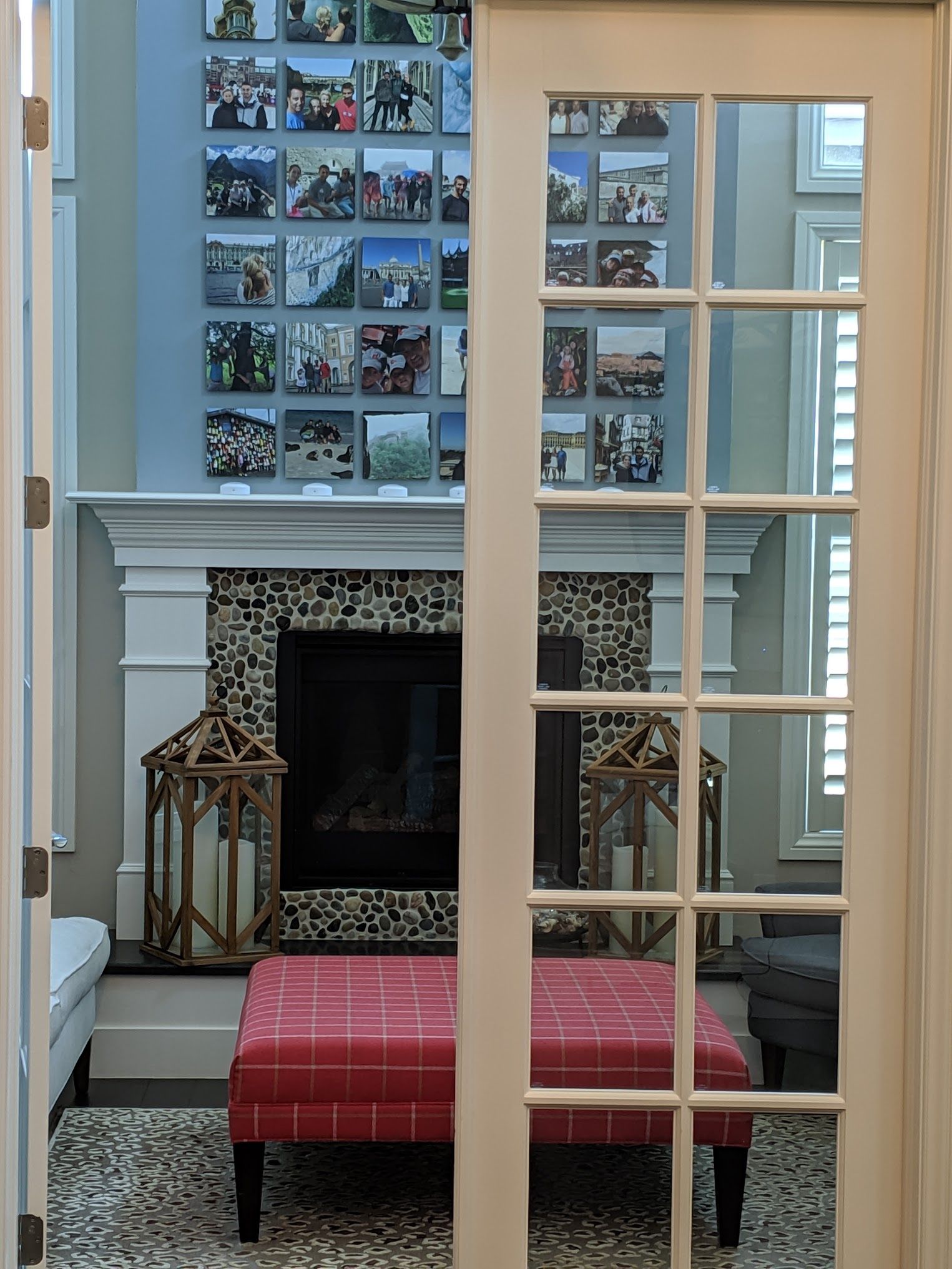 A living room with a fireplace, photos on the wall, and a red plaid ottoman, viewed through a glass door.