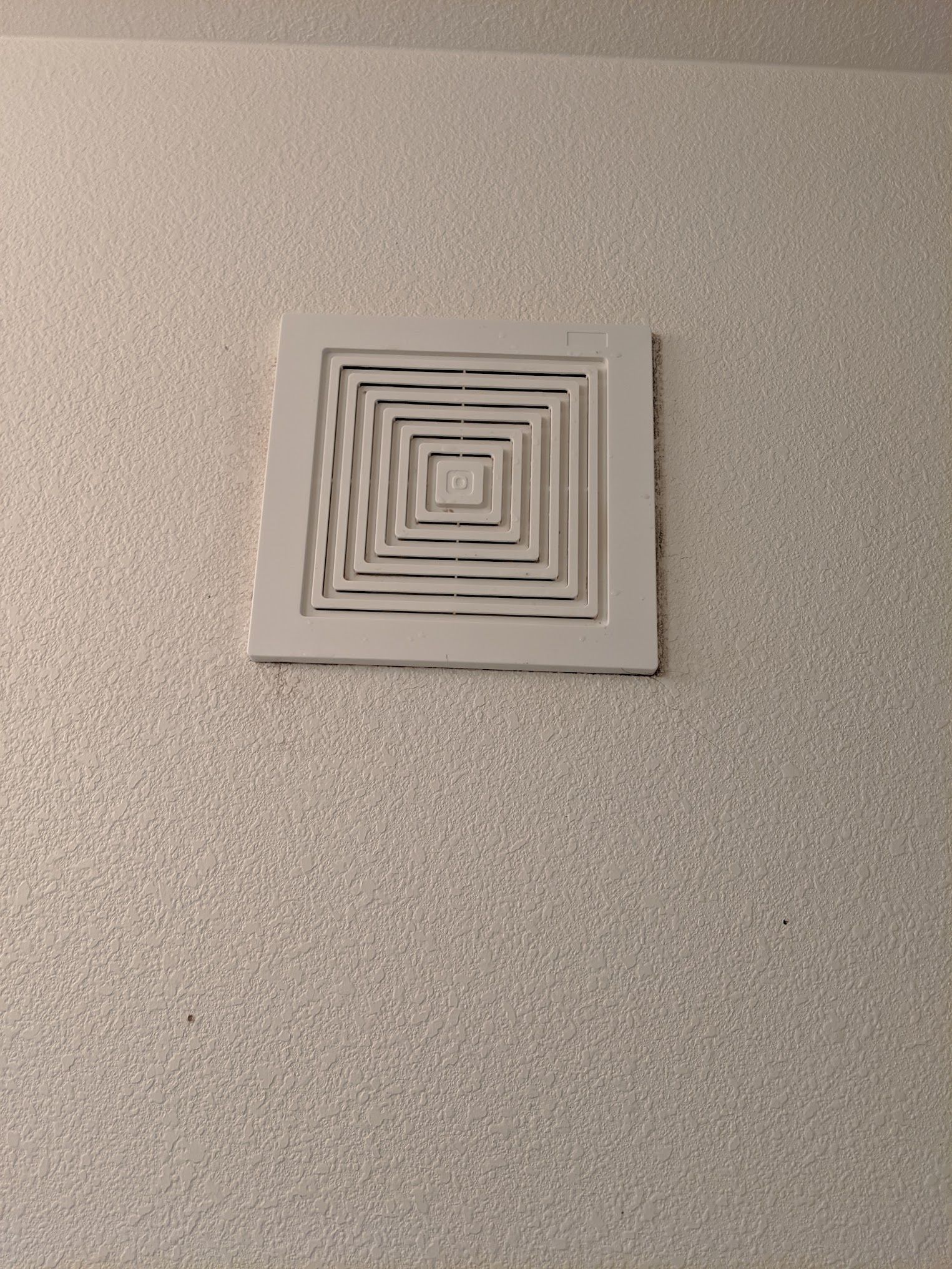 White, square vent cover with concentric square louvers on a textured wall.