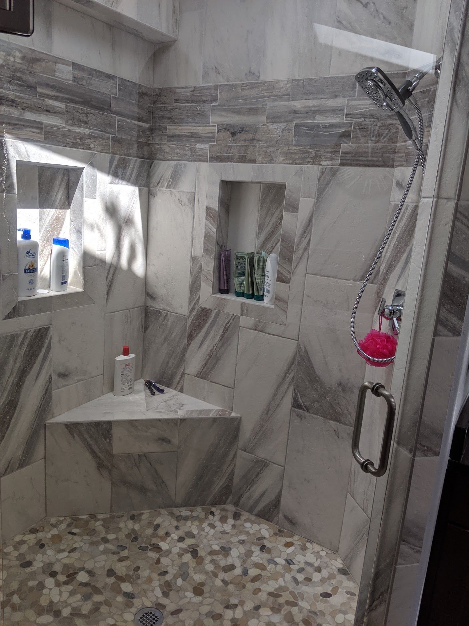 A modern bathroom shower with gray marble and stone tile walls, pebble floor, and built-in shelves.