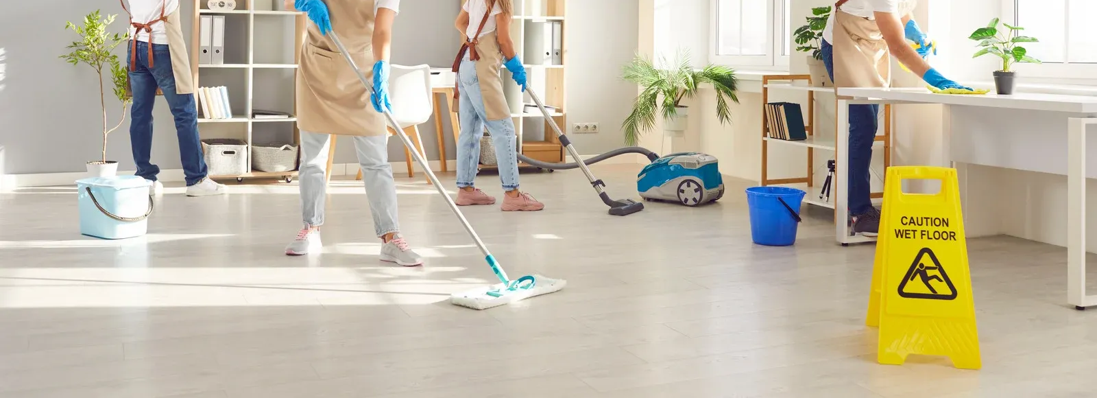 Four cleaners cleaning an office with mops, vacuum, and wet floor sign present.