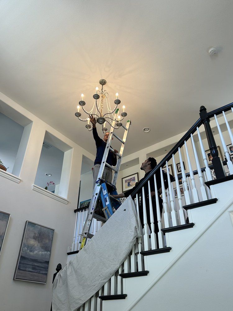 A man on a ladder is fixing a chandelier in a living room.