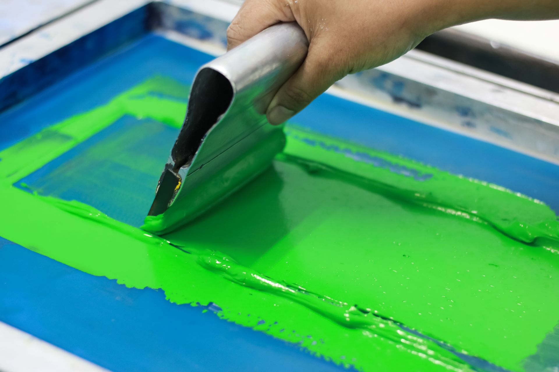 A hand uses a squeegee to spread bright green ink across a blue screen printing frame.