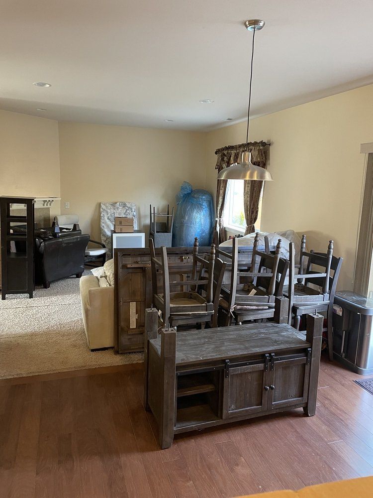 A room with furniture stacked, including a cabinet, chairs, and a blue bag.