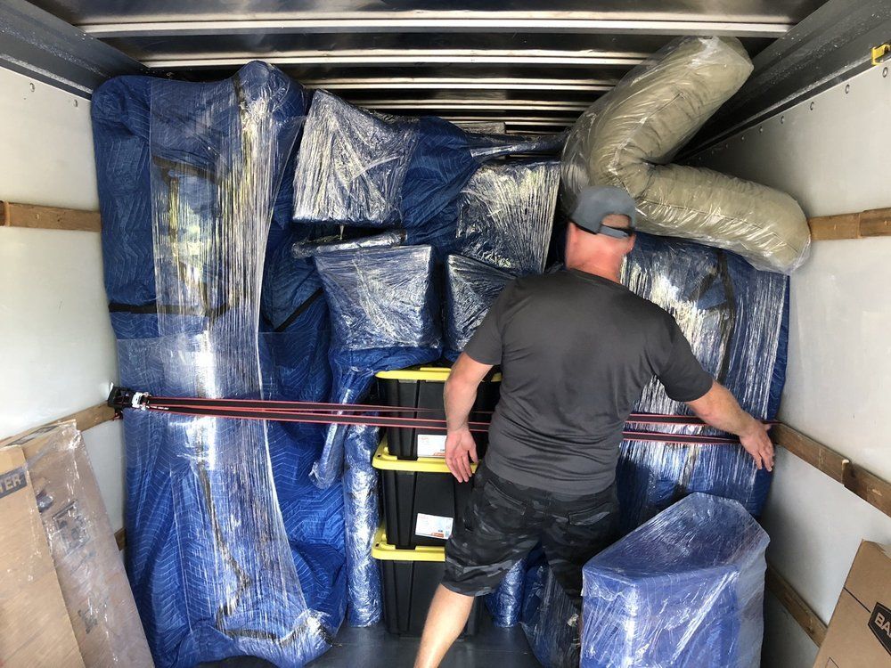 Man securing wrapped furniture and boxes in a moving truck with a cargo strap.