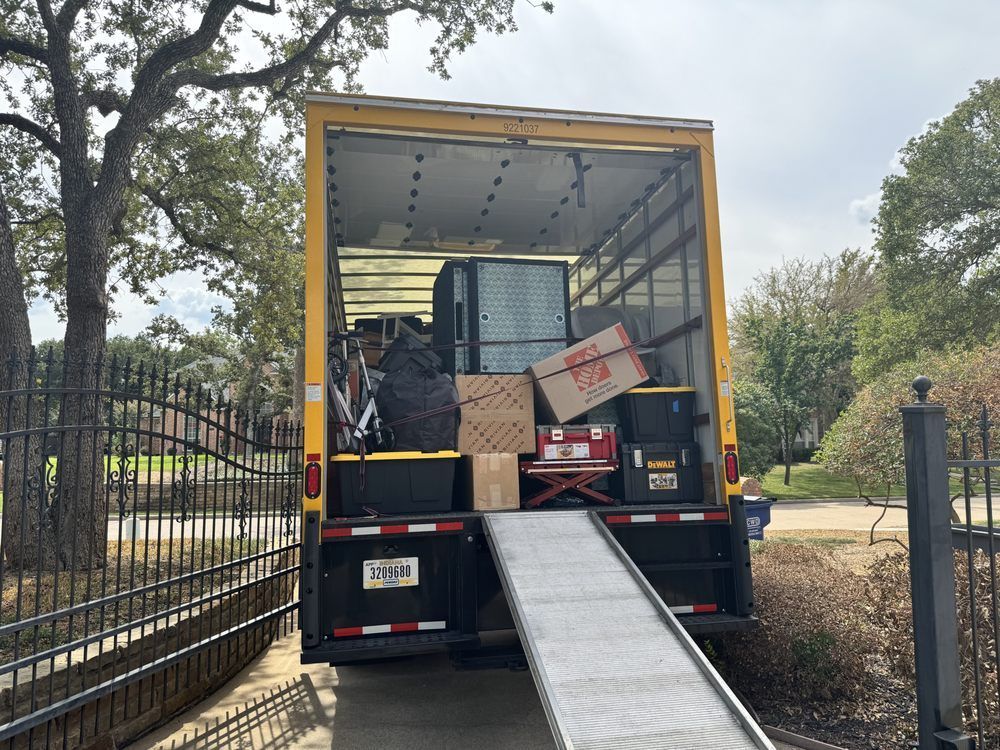 A moving truck with ramp extended, filled with boxes and furniture, parked in a driveway with a black gate.