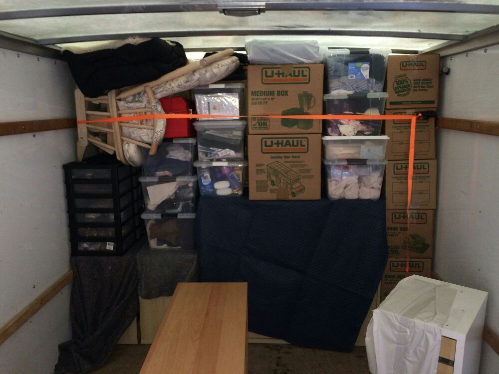 A truck interior filled with boxes, furniture, and bins, secured with an orange strap, ready for transport.