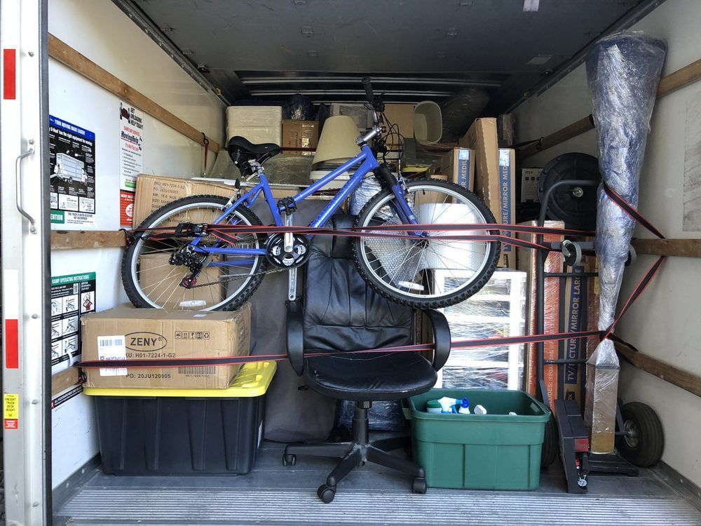Inside a moving truck, a blue bike, boxes, a chair, and other items are secured with straps.