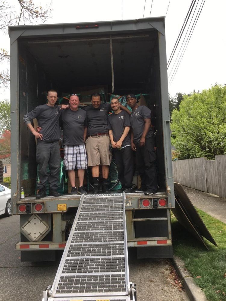 Five movers pose in the open back of a moving truck with a ramp extended, on a residential street.