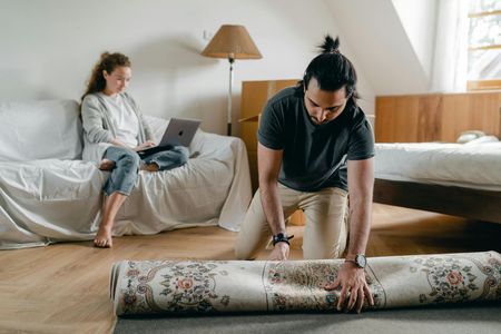 A person rolling out a rug while another person works on a laptop on a couch. Room interior.
