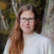 Mari Rantanen wearing glasses and a white sweater is standing in front of a tree.