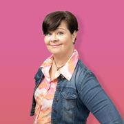 Susa Laine wearing a denim jacket and a colorful shirt is standing in front of a pink background.