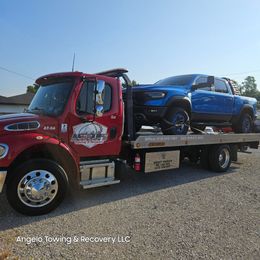Red tow truck with a blue pickup truck on the flatbed, under a clear sky. Angelo Towing & Recovery LLC.