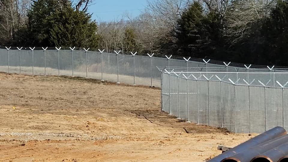 A chain link fence is surrounding a dirt field with trees in the background.