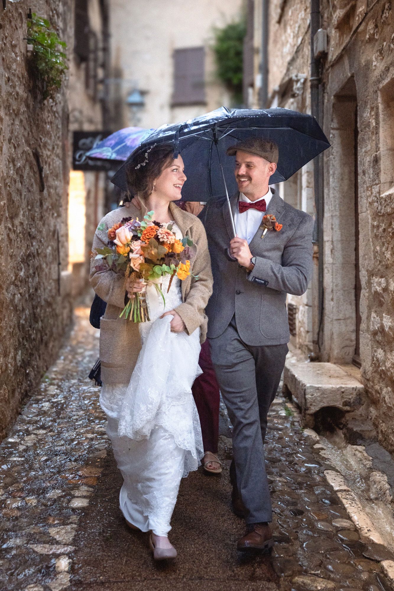 A bride and groom walking under an umbrella through St Paul de Vence in the rain on their wedding day.