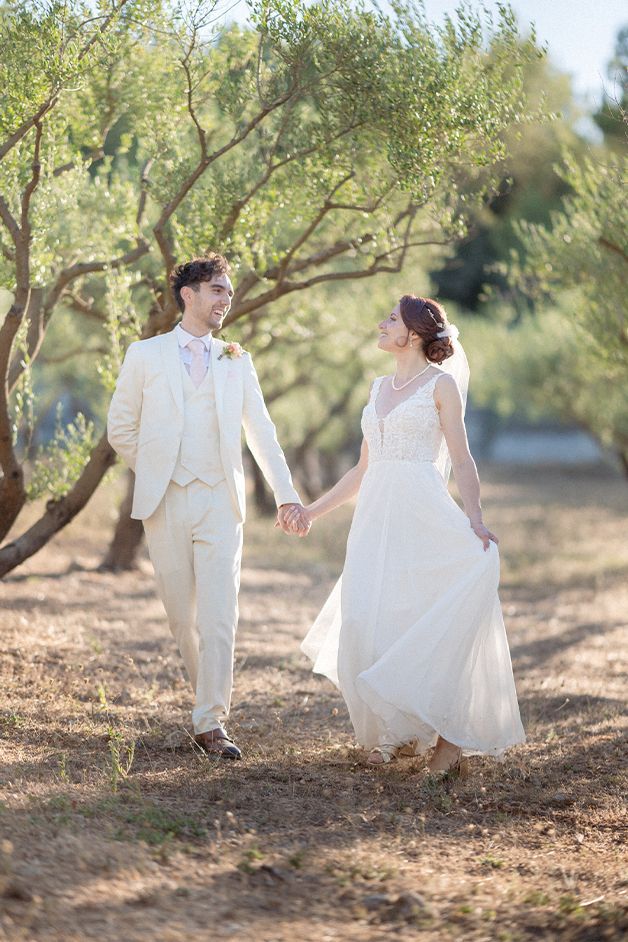 A bride and groom smile and laugh as they walk through an olive grove at their wedding in the South of France.