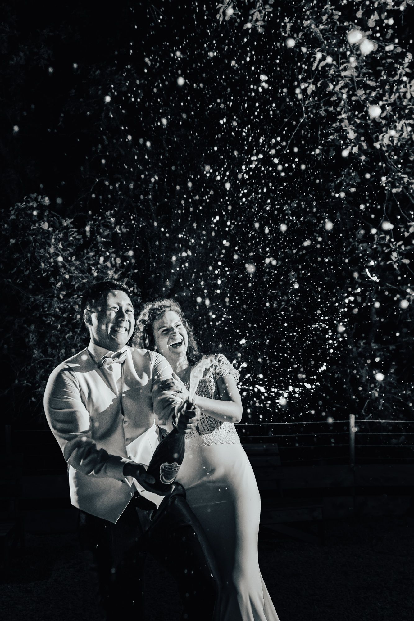 A champagne bottle exploding at a wedding lit by flash while the couple laugh.