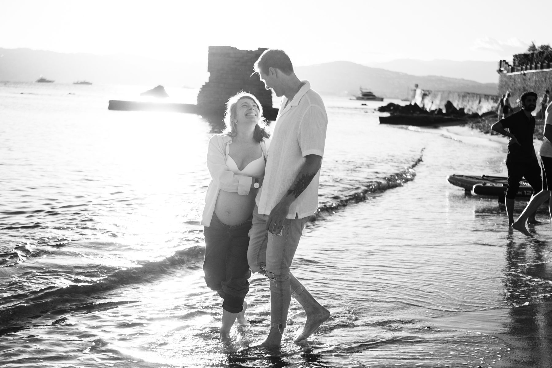 A couple laugh as they walk along a beach during a maternity photography session.