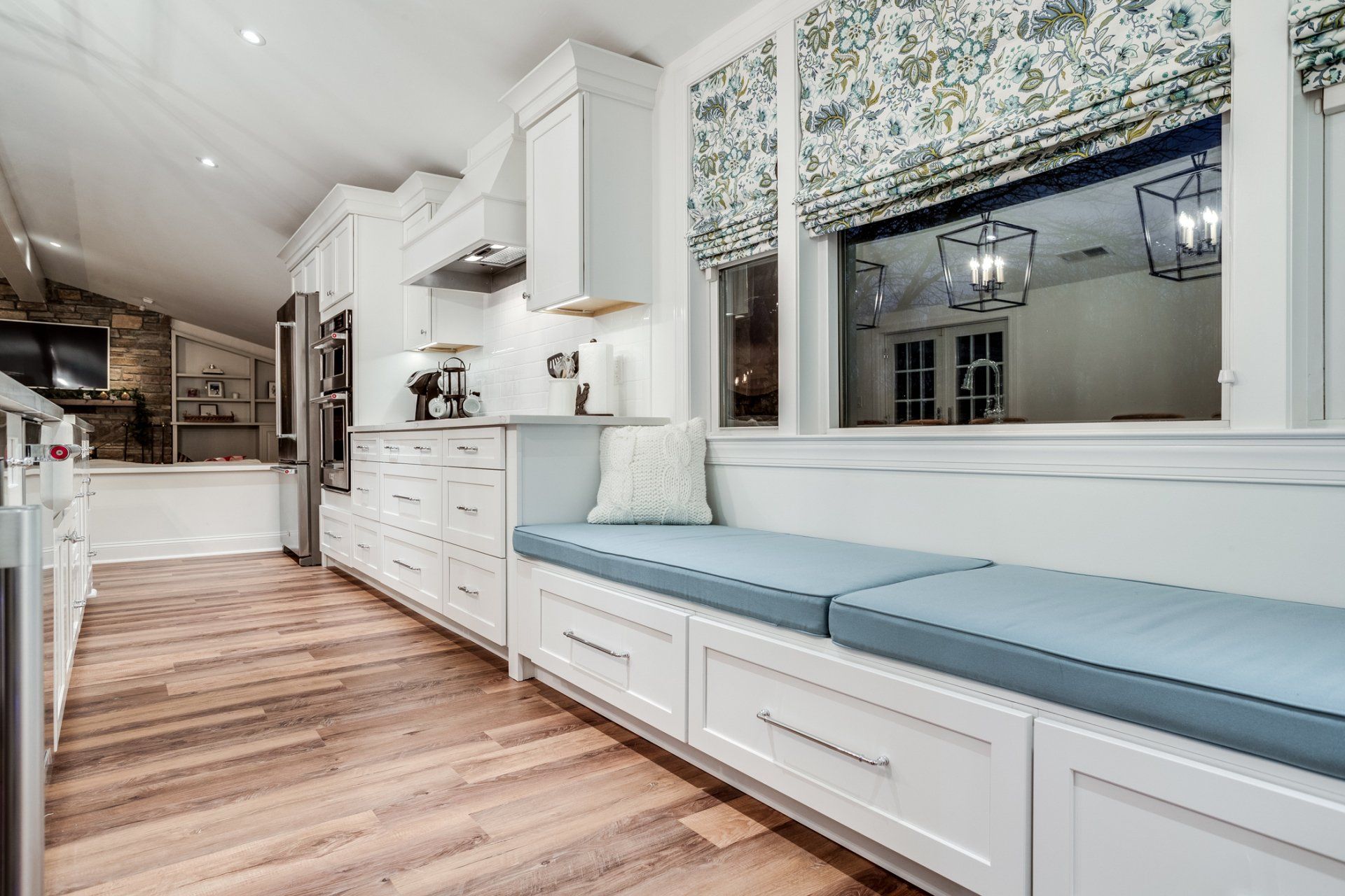 A kitchen with white cabinets , hardwood floors , a bench and a window.