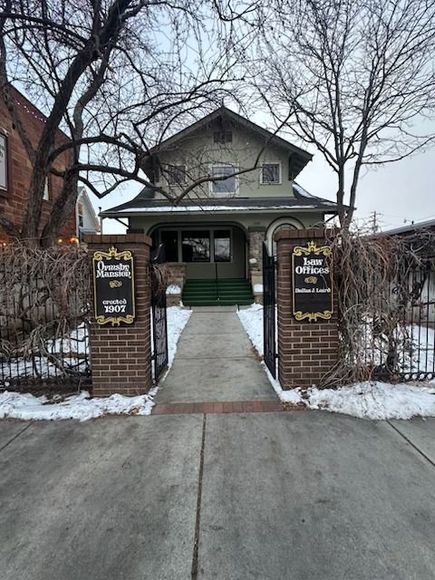 A path leads to an old house with a green porch between two brick pillars holding signs for
