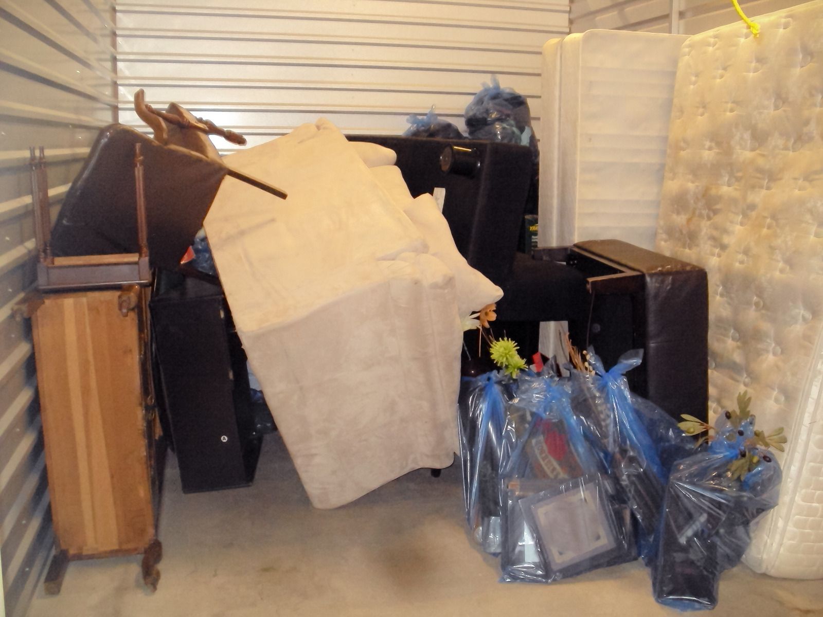 A storage unit packed with a mattress, furniture covered by a blanket, and several blue plastic bags on the floor.